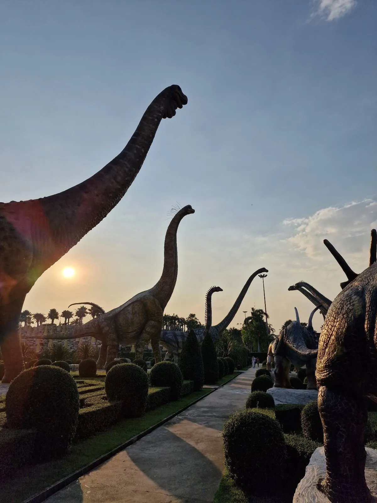 Silhouettes of dinosaur statues, including long-necked sauropods, stand against a sunset sky. The scene is set in a landscaped garden with a clear pathway and neatly trimmed bushes.