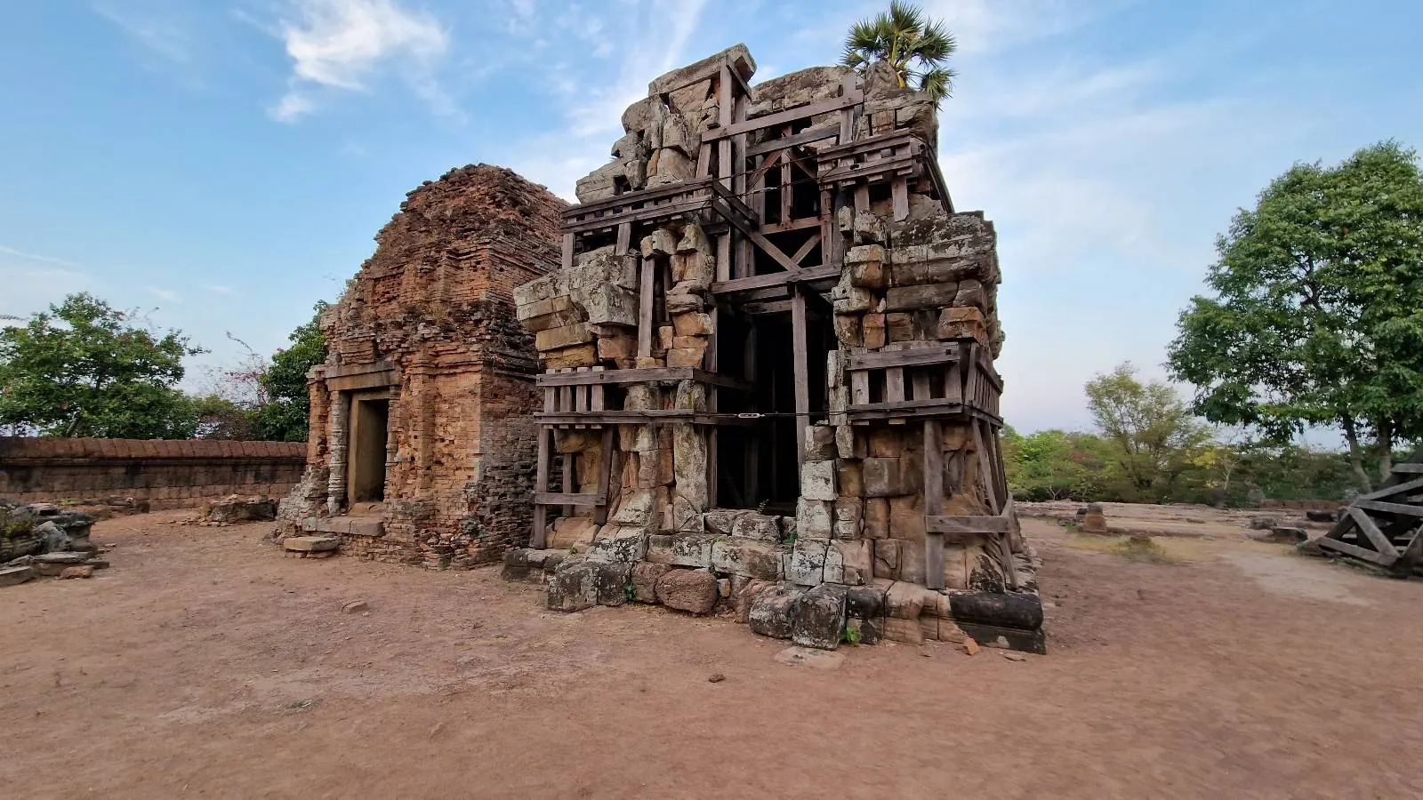Ancient stone temple ruins with intricate carvings and wooden supports, surrounded by trees and an open dirt area under a partly cloudy sky.