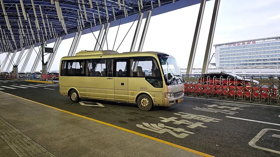 A beige minibus parked at a modern, open-air terminal with blue canopies and glass. Japanese characters on the road. Building in the background.