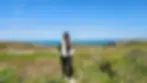 A woman in a white jacket and black pants stands smiling on a coastal grassland under a clear blue sky.