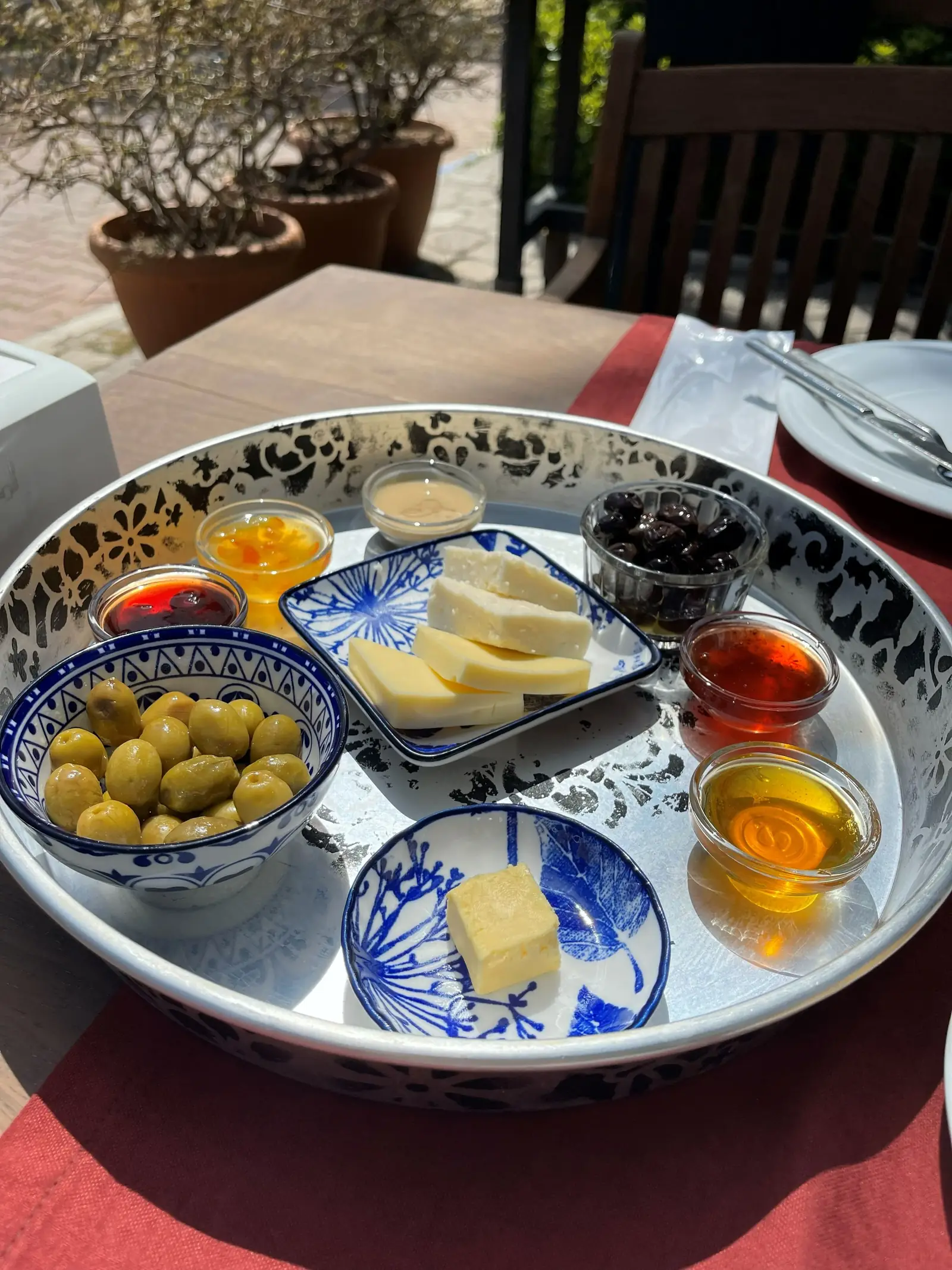 A large round tray with bowls of green olives, red and orange sauces, cheese slices, and a center block of butter on a table with red tablecloth.