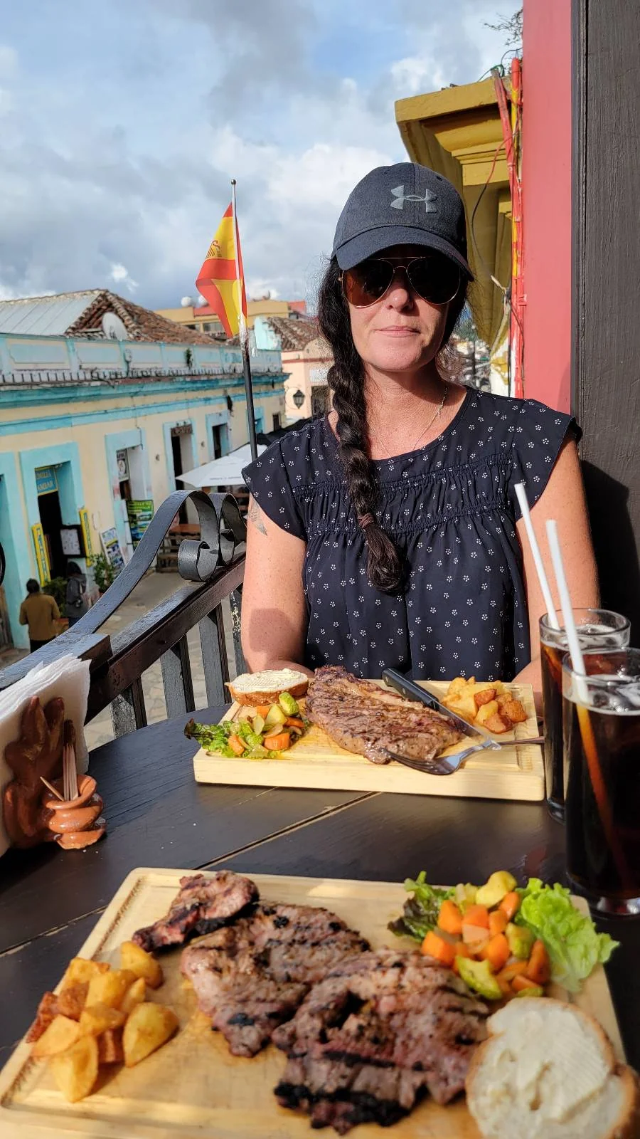 A woman in a blue cap sits at an outdoor table with two wooden boards of food. Each board has grilled meat, vegetable garnish, and diced potatoes. A glass of dark soda with a straw is beside her. Colorful buildings line the street in the background.
