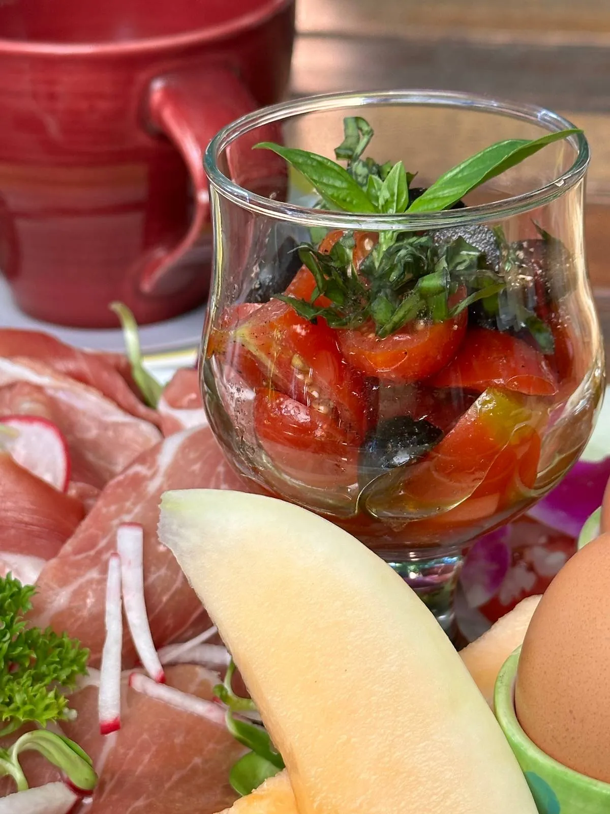 A glass cup filled with a tomato and basil salad is surrounded by assorted food items, including slices of prosciutto, melon, radishes, and a brown egg. A red mug is partially visible in the background.
