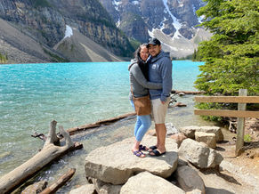 A couple embraces on a rock by the azure blue lake in Banff National Park, with tree-lined shores and snowy mountains in the background. They are both wearing casual clothing and look happy. The sky is clear, and logs and rocks are scattered near the water's edge.