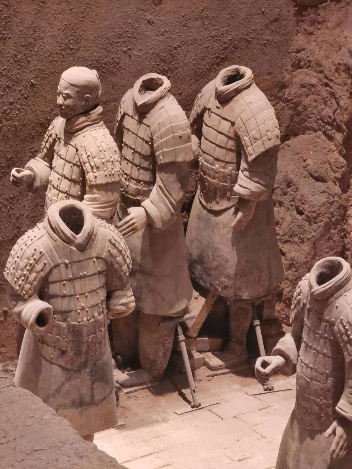 Four headless terracotta warrior statues stand in an excavation site, wearing detailed armor and robes, surrounded by earthen walls.