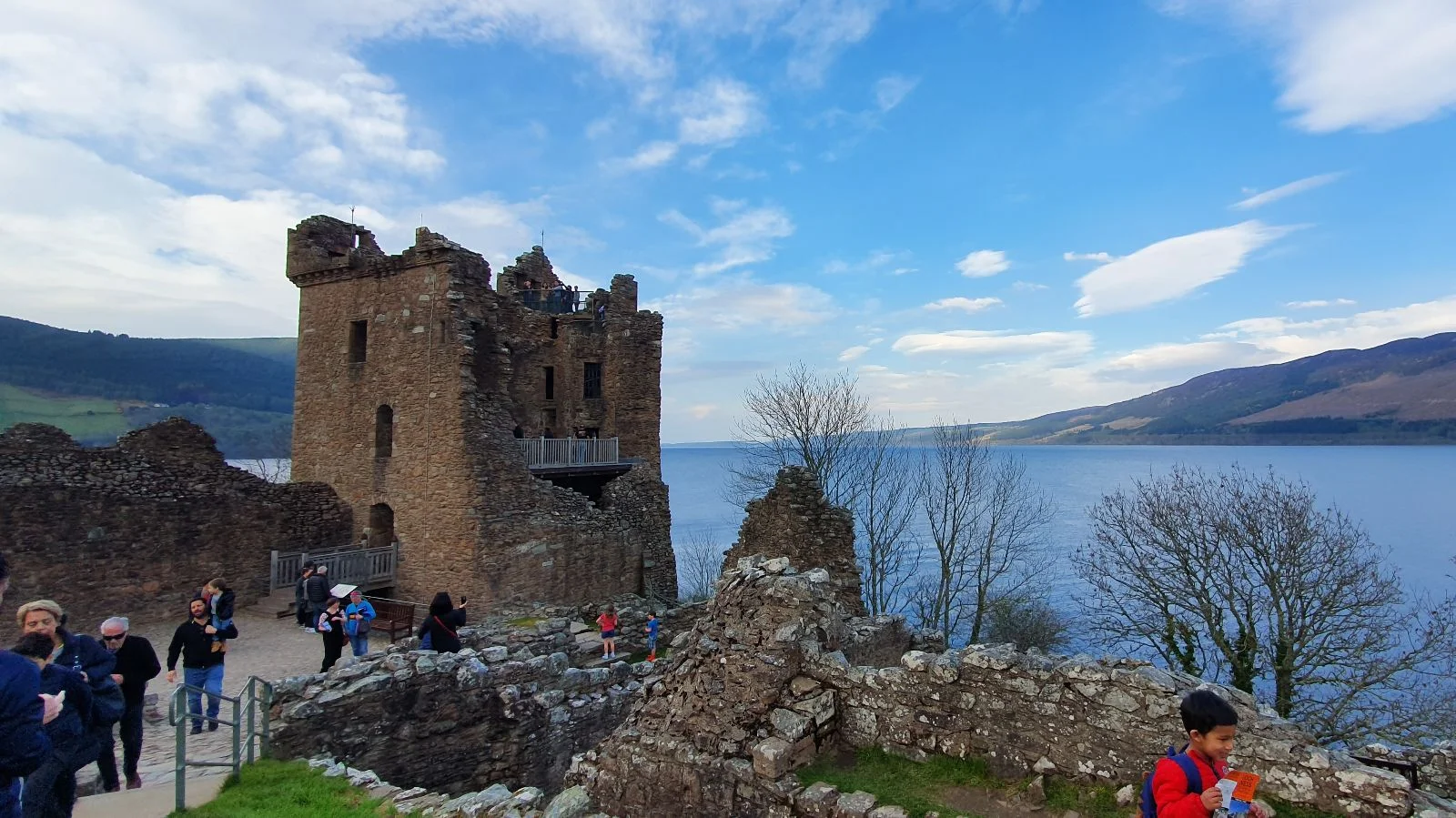 Tower house at Urquhart Castle overlooking Loch Ness, with visitors walking along the path and ruins in the foreground.