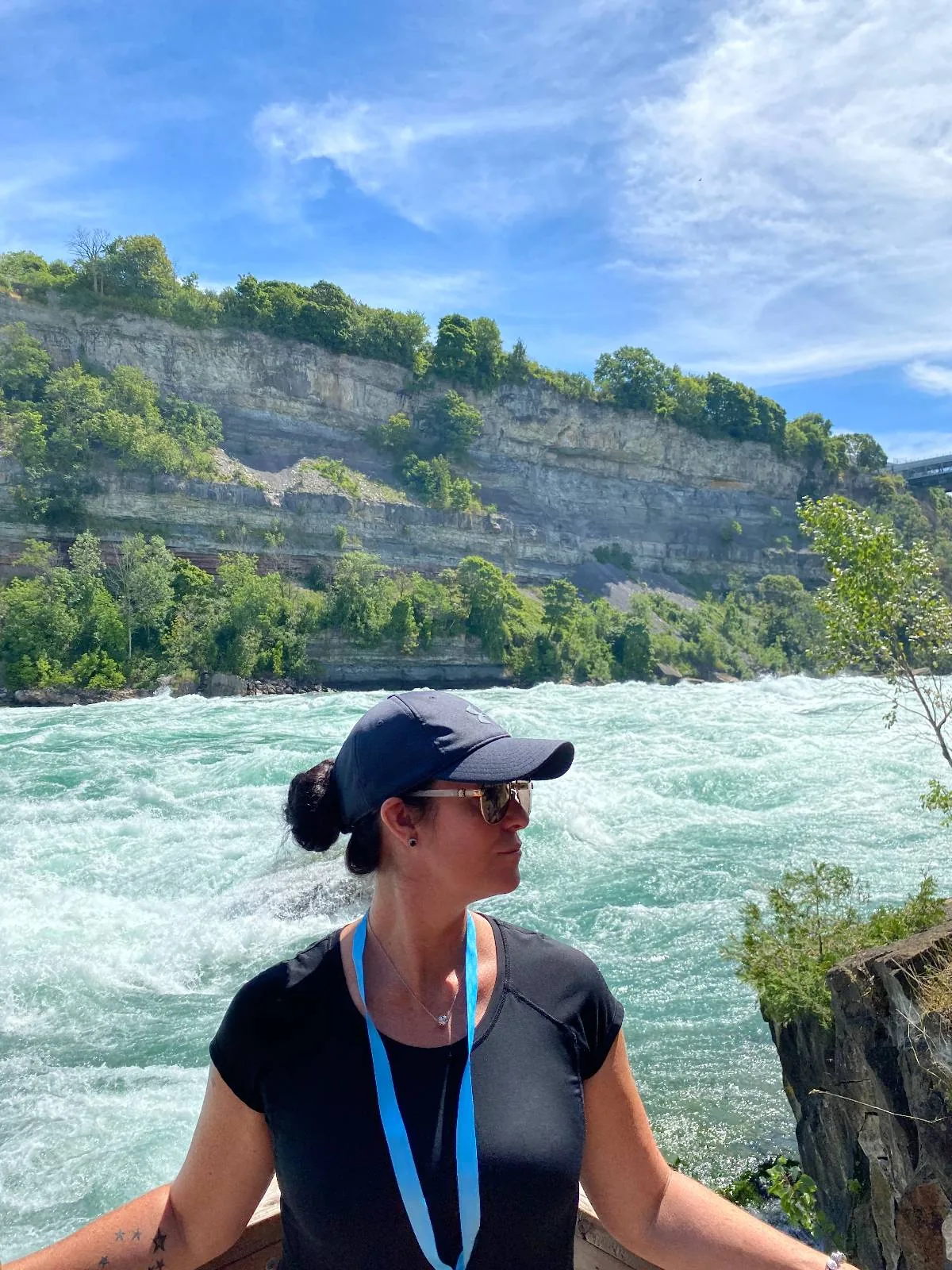 A person stands in front of a fast-flowing river with a rocky cliff in the background.