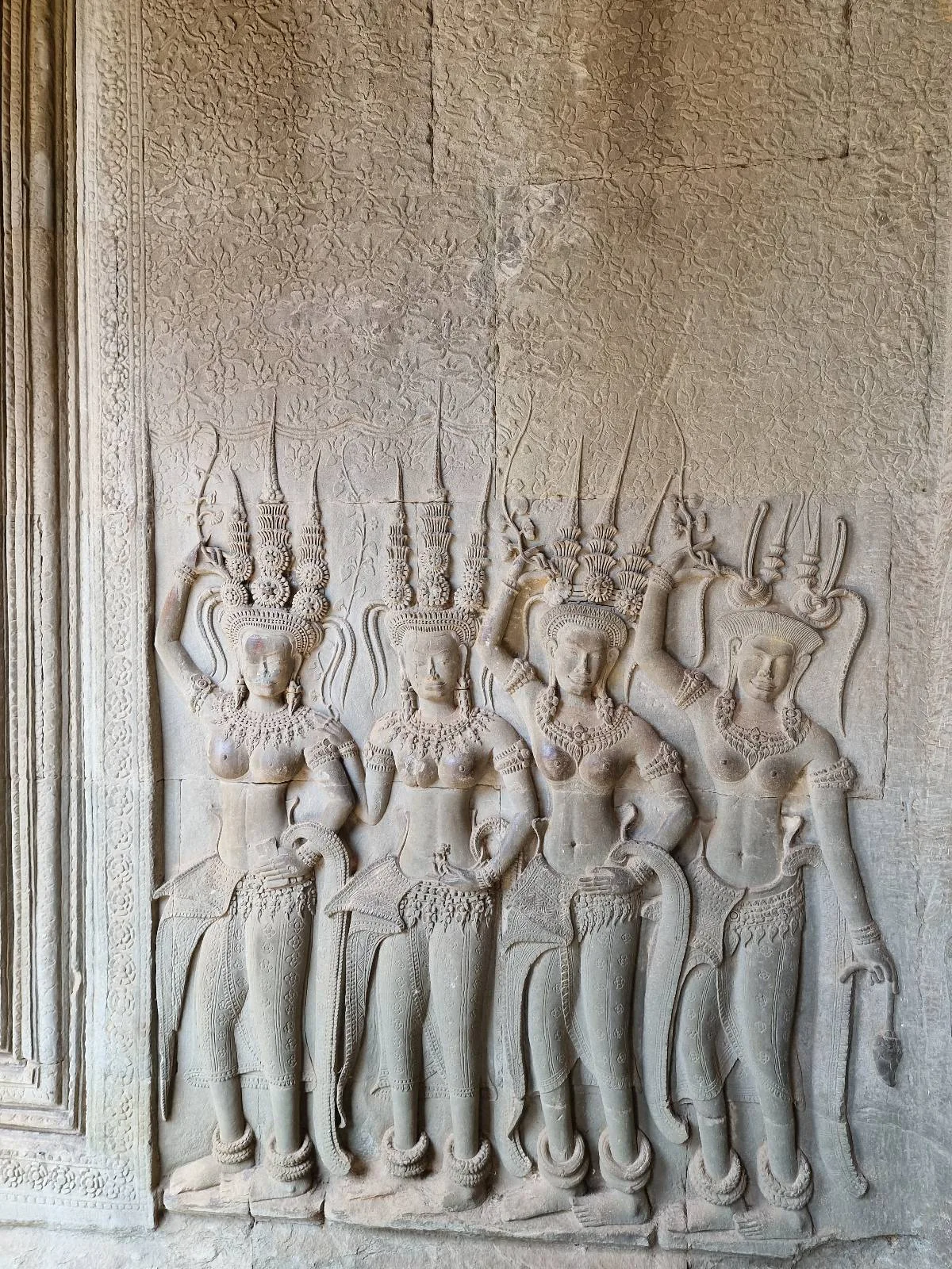 Stone carving of four female figures in traditional attire with elaborate headdresses, standing side by side on a wall at Angkor Wat, each holding decorative poses with their hands raised.