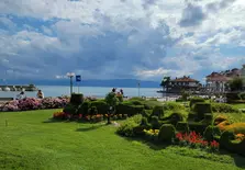 A scenic lakeside view with colorful flower beds in the foreground, showcasing orange, yellow, and pink blooms. Two people enjoy the seating areas by the beautiful lake, with a house and cloudy sky in the background.