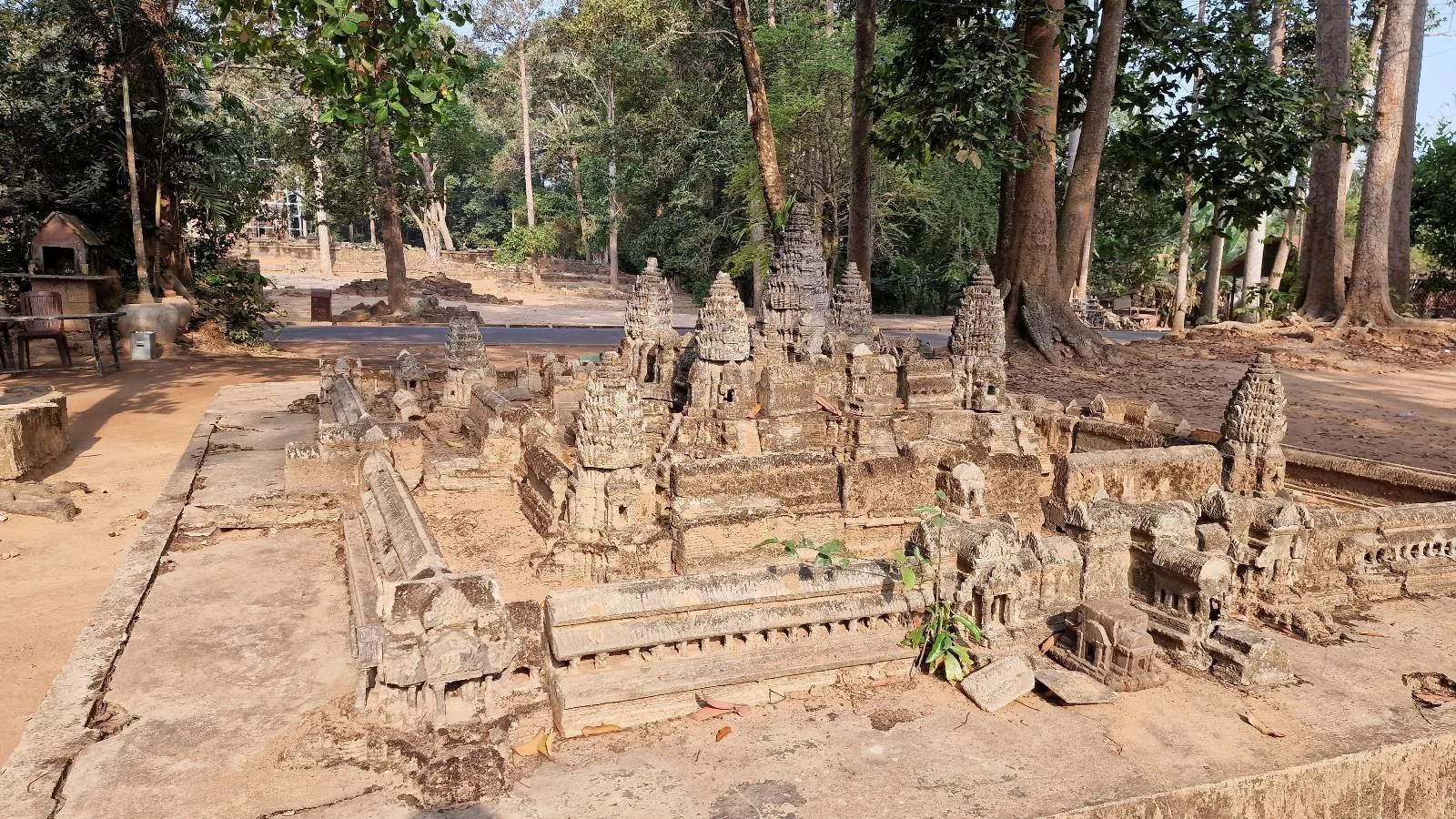 A detailed stone model of Angkor Wat sits outdoors surrounded by trees, with a dirt path and lush forest visible in the background.