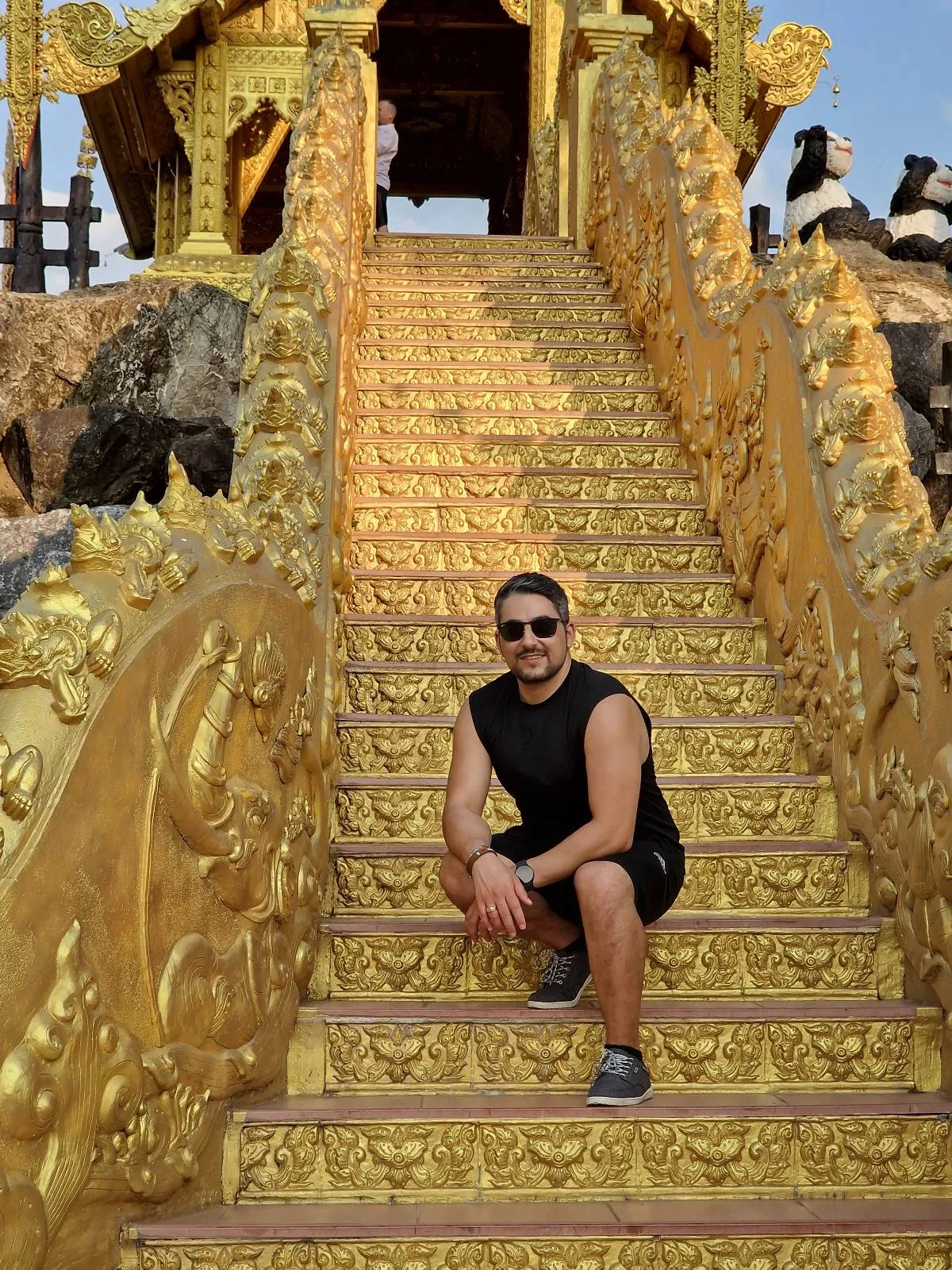 A man wearing sunglasses and a sleeveless black shirt crouches on ornately decorated golden stairs with intricate carvings. The structure is part of a richly adorned building. The sky is clear and sunny.