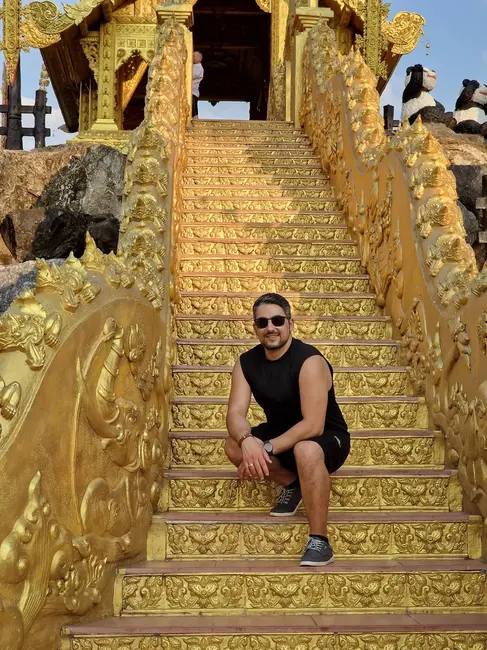 A man wearing sunglasses and a sleeveless black shirt crouches on ornately decorated golden stairs with intricate carvings. The structure is part of a richly adorned building. The sky is clear and sunny.