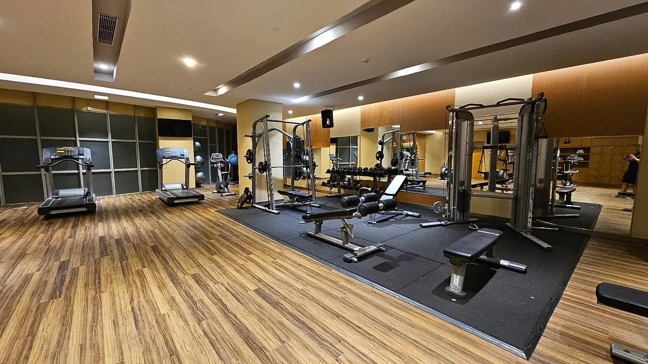 A spacious, modern gym with wooden floors and mirrored walls. It features treadmills, weight benches, and various exercise machines. The room is well-lit, and the equipment is neatly arranged.