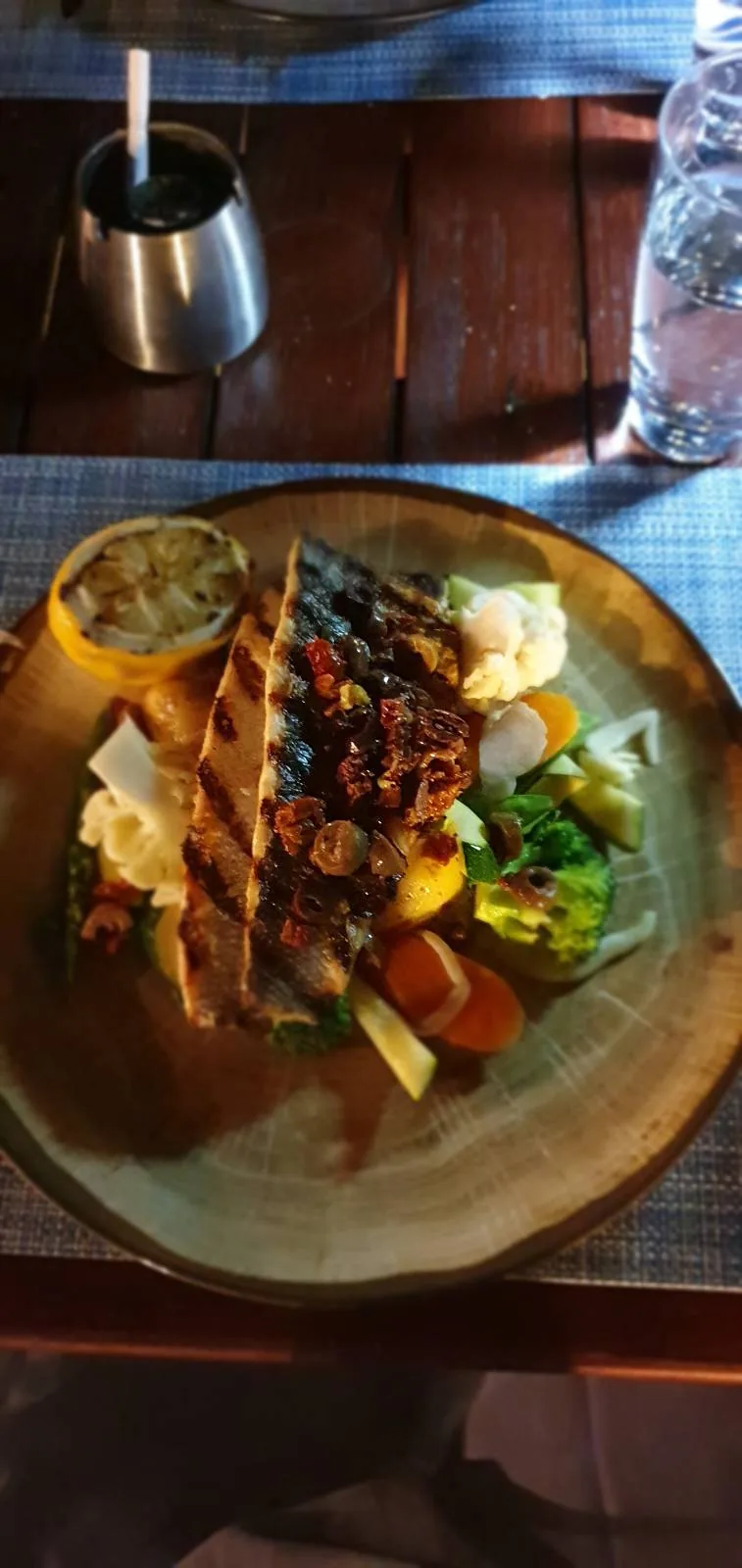Grilled fish fillet topped with spices, served on a round plate with a side of mixed vegetables and a lemon slice. The dish is set on a wooden table with a blue placemat, accompanied by a glass and a small metal container.