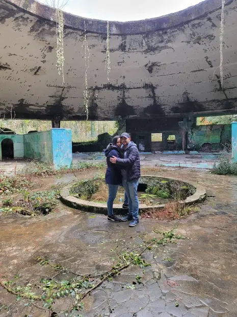 A couple embraces in the center of an abandoned, circular, open-roofed structure in Tskaltubo, overgrown with plants and covered in graffiti—natural light streams down, recalling the haunting beauty of its Soviet-era sanatoriums.