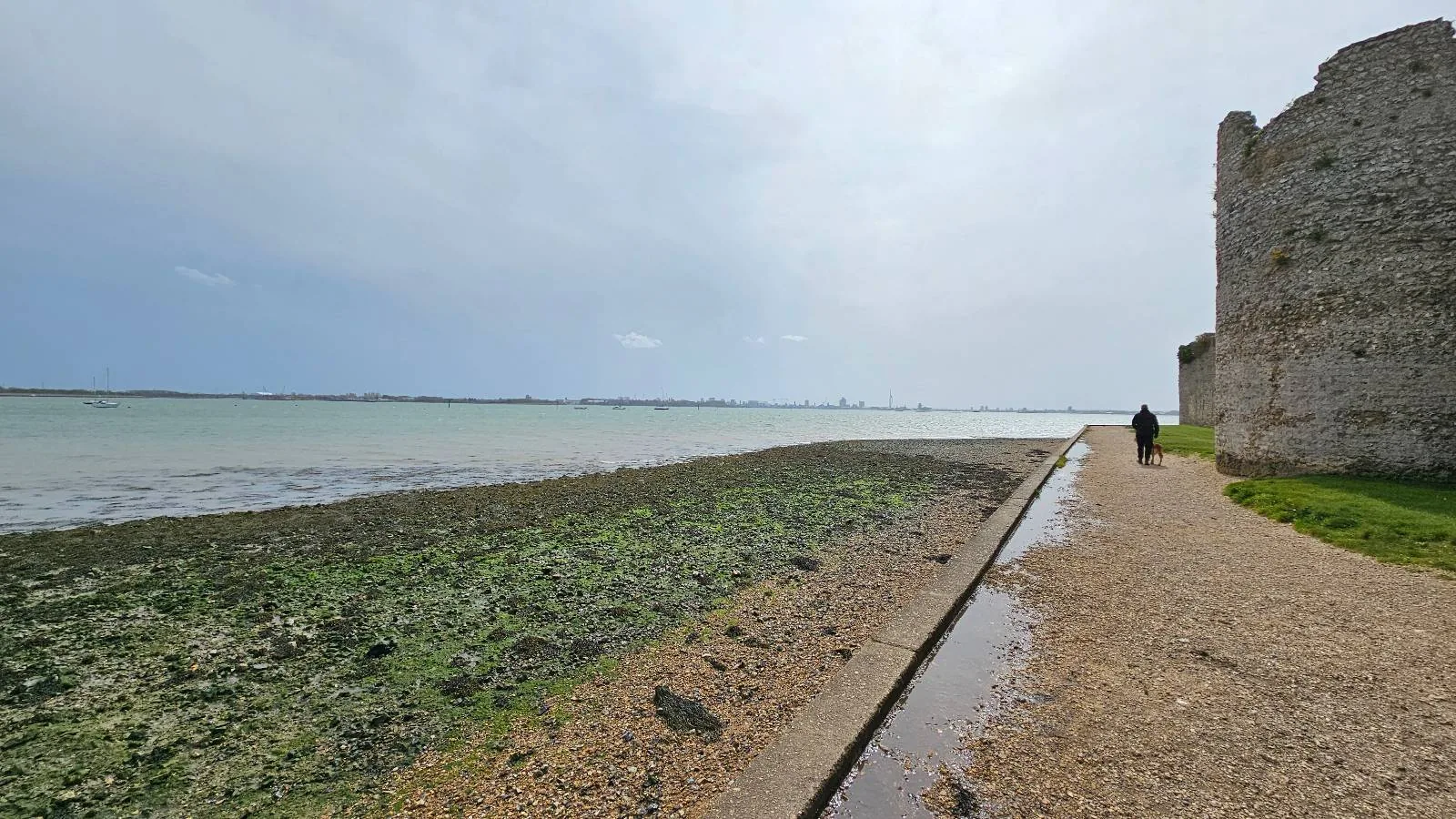 A wide view of a coastal pathway beside an ancient stone wall with two people walking, overlooking a calm sea under a cloudy sky.