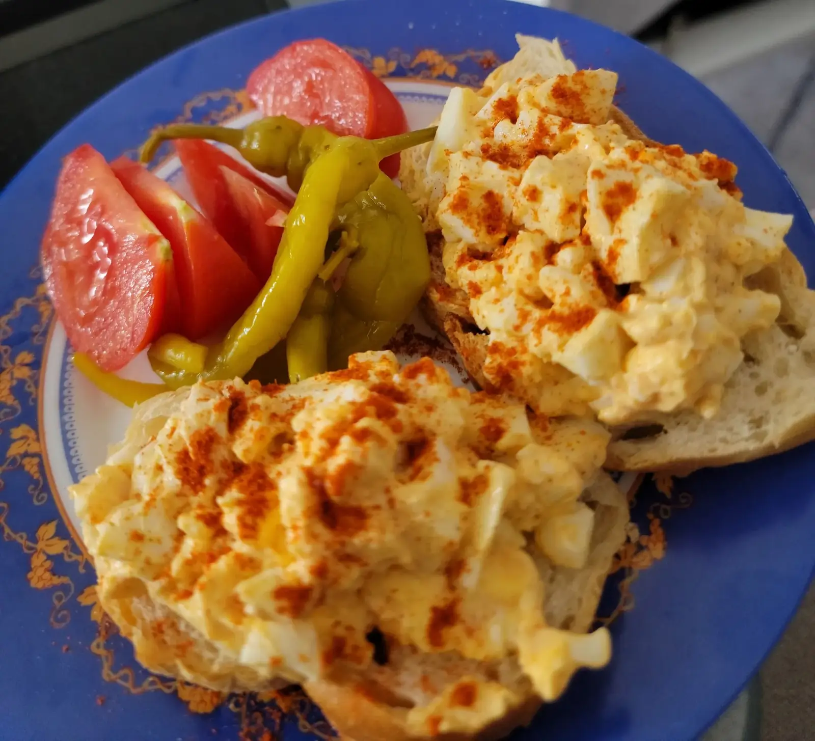 Plate with egg salad on bread, sprinkled with paprika, served with tomato slices and yellow peppers.