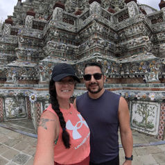 A woman in a pink tank top and black cap stands next to a man in sunglasses and a dark sleeveless shirt. Embracing slow travel, they smile in front of an ornate stone temple with intricate carvings, embodying the essence of digital nomads exploring the world.