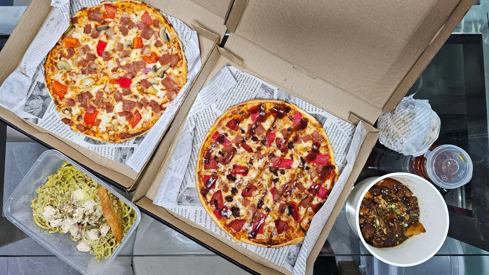 Two open pizza boxes with different pizzas, a container of pasta with cauliflower, and a bowl of stew or curry are arranged on a glass table. Disposable sauce containers are also visible.