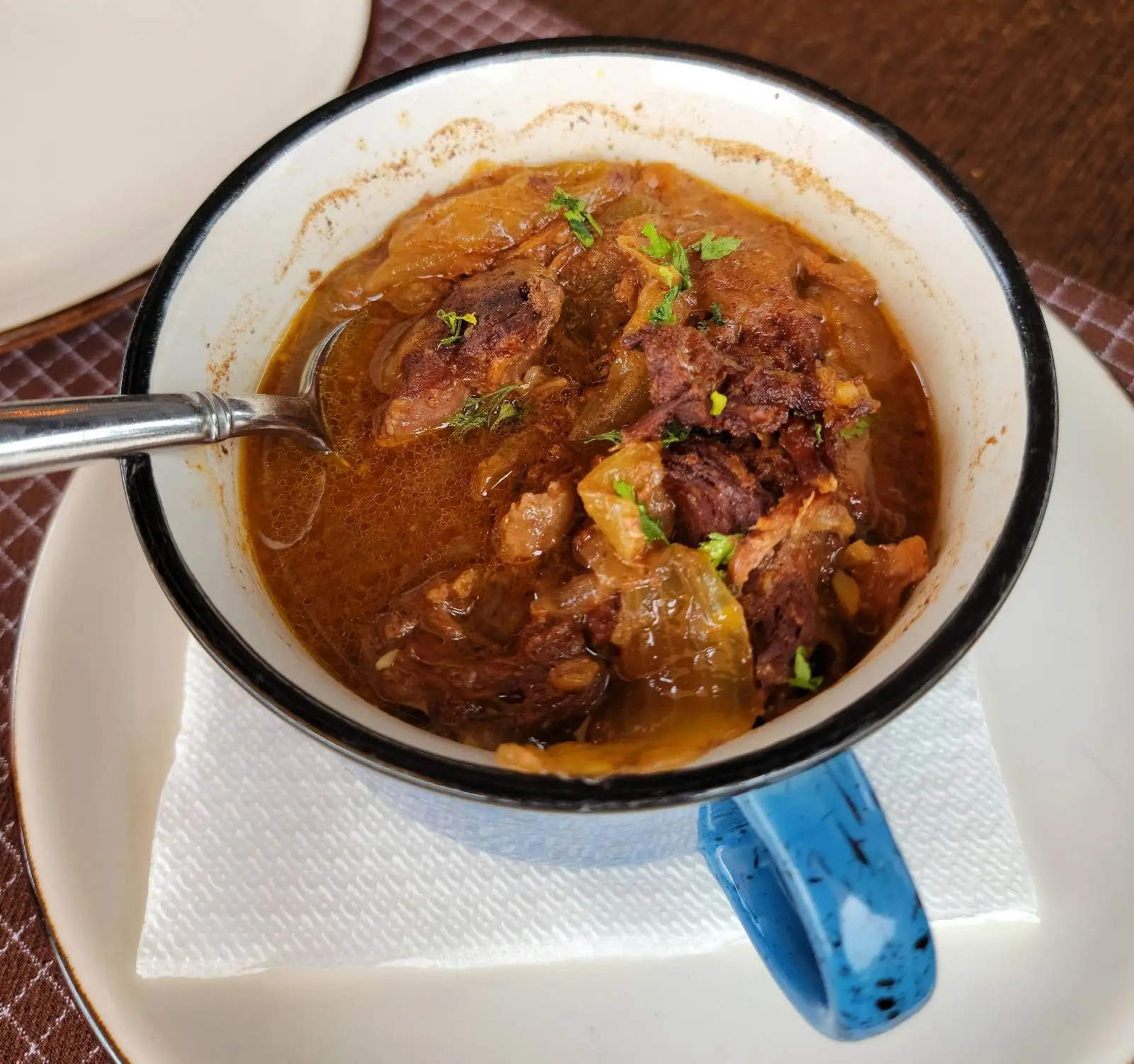 A mug filled with hearty beef stew, garnished with chopped herbs. The stew contains chunks of meat and vegetables in a rich, brown broth. A spoon rests inside the mug, which is set on a white napkin and plate.