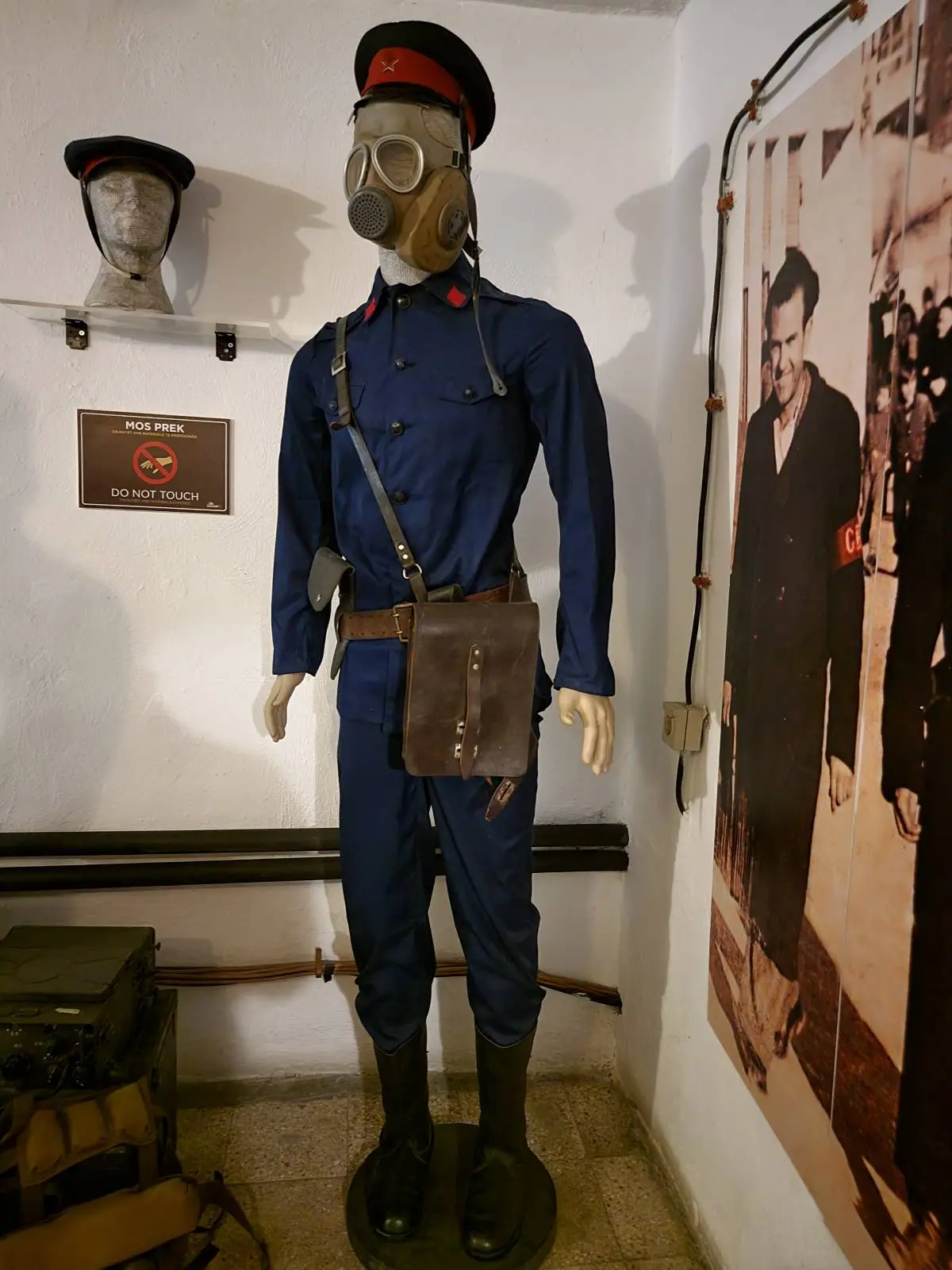 A mannequin dressed in a vintage military or paramilitary uniform with a gas mask, blue attire, and a red-trimmed cap. It has a satchel slung across the shoulder. Nearby, a black-and-white photograph and a mounted gas mask on the wall are visible.