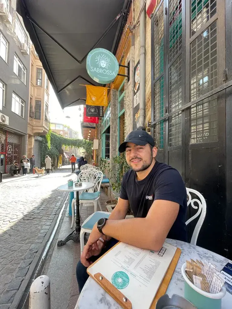 A man in a black t-shirt and cap sits at a small outdoor café table on a cobblestone street, smiling at the camera. There is a menu and a plate on the table, with buildings and more tables in the background.