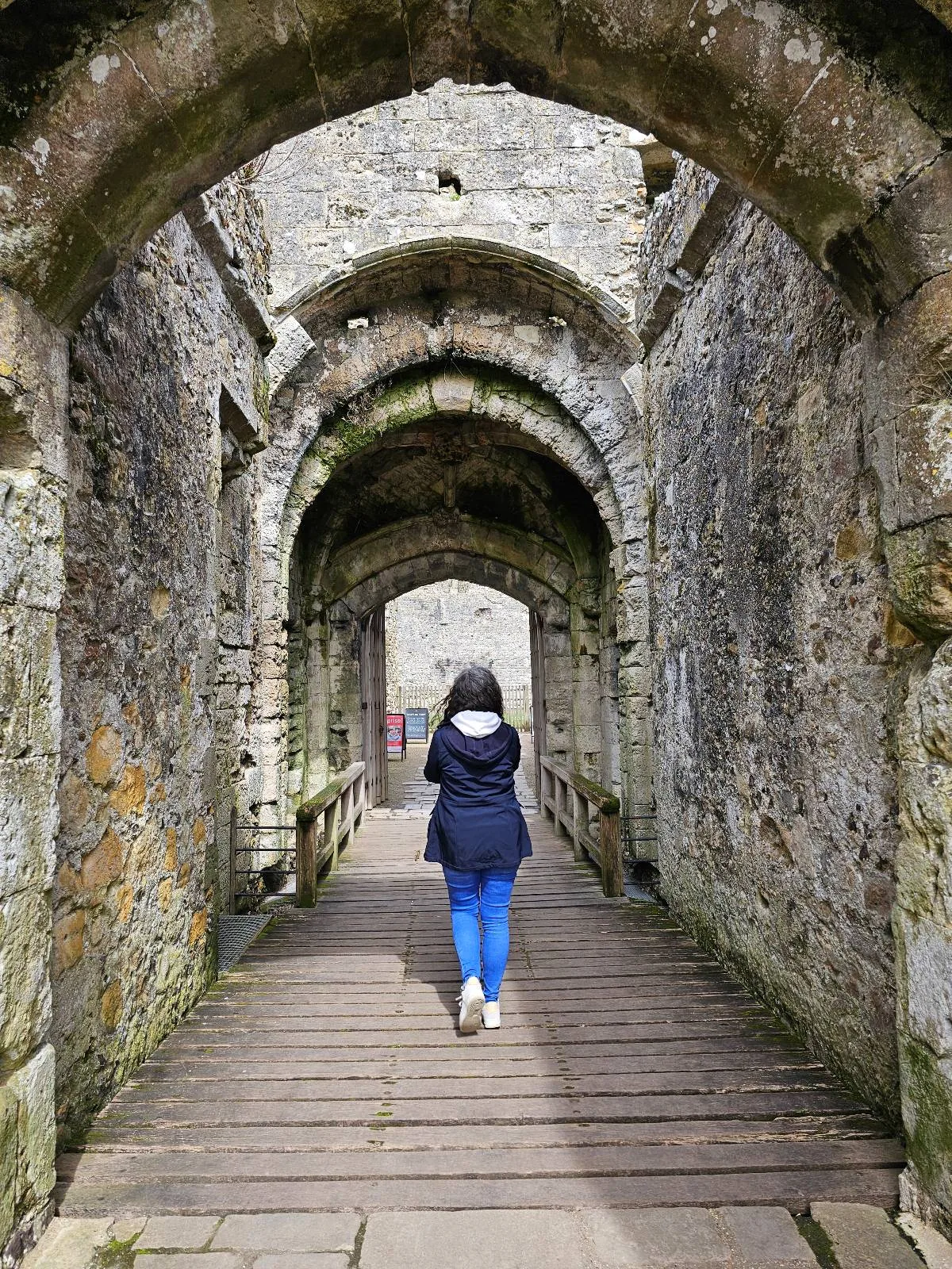 A person in a blue jacket and jeans walks through a stone archway in an old, weathered building, with sunlight illuminating the path ahead.
