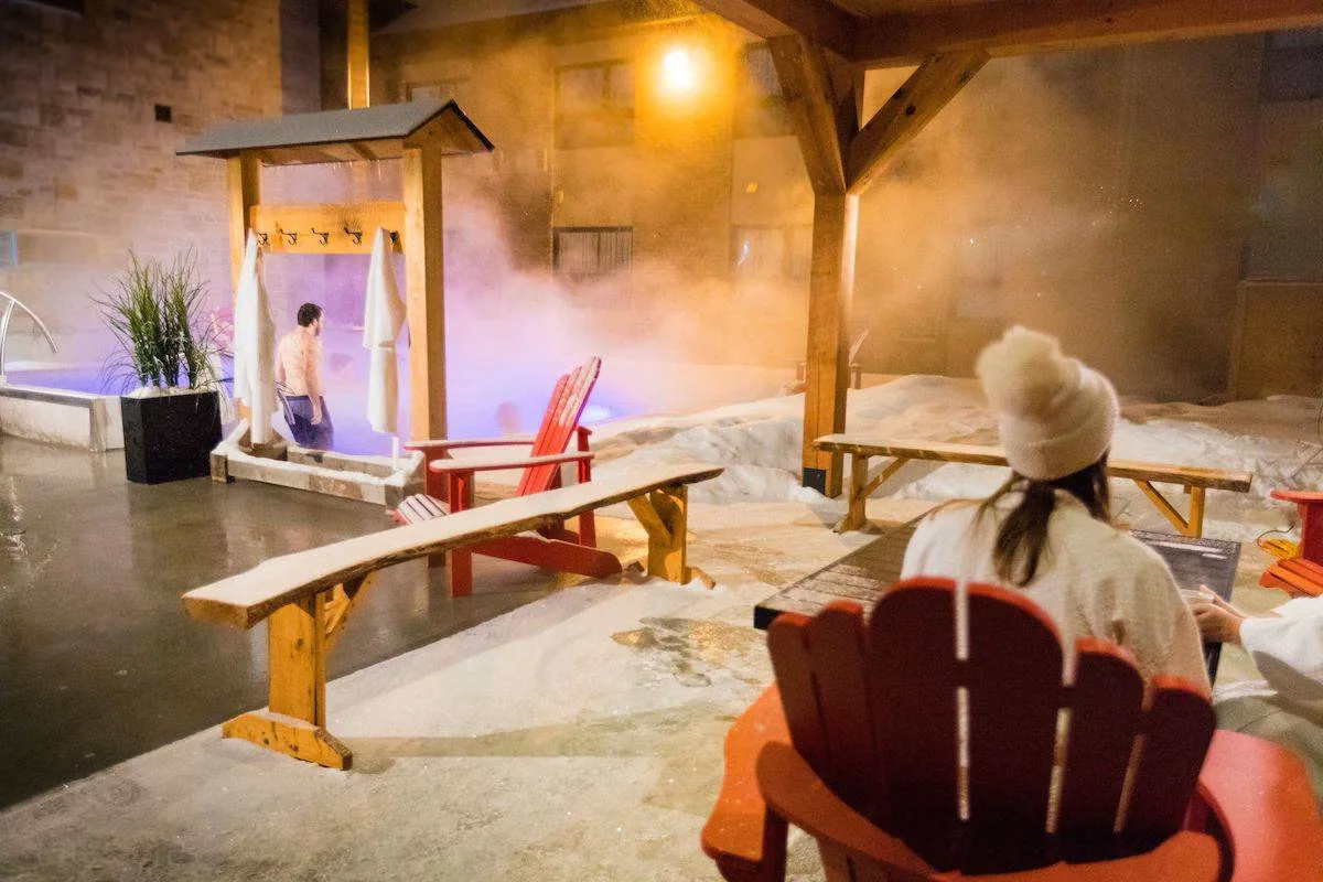 A person in a robe and hat sits on a red Adirondack chair near a snowy pool area with steam rising. Another person stands under a wooden shower structure. Warm lighting and wooden beams give a cozy ambiance.