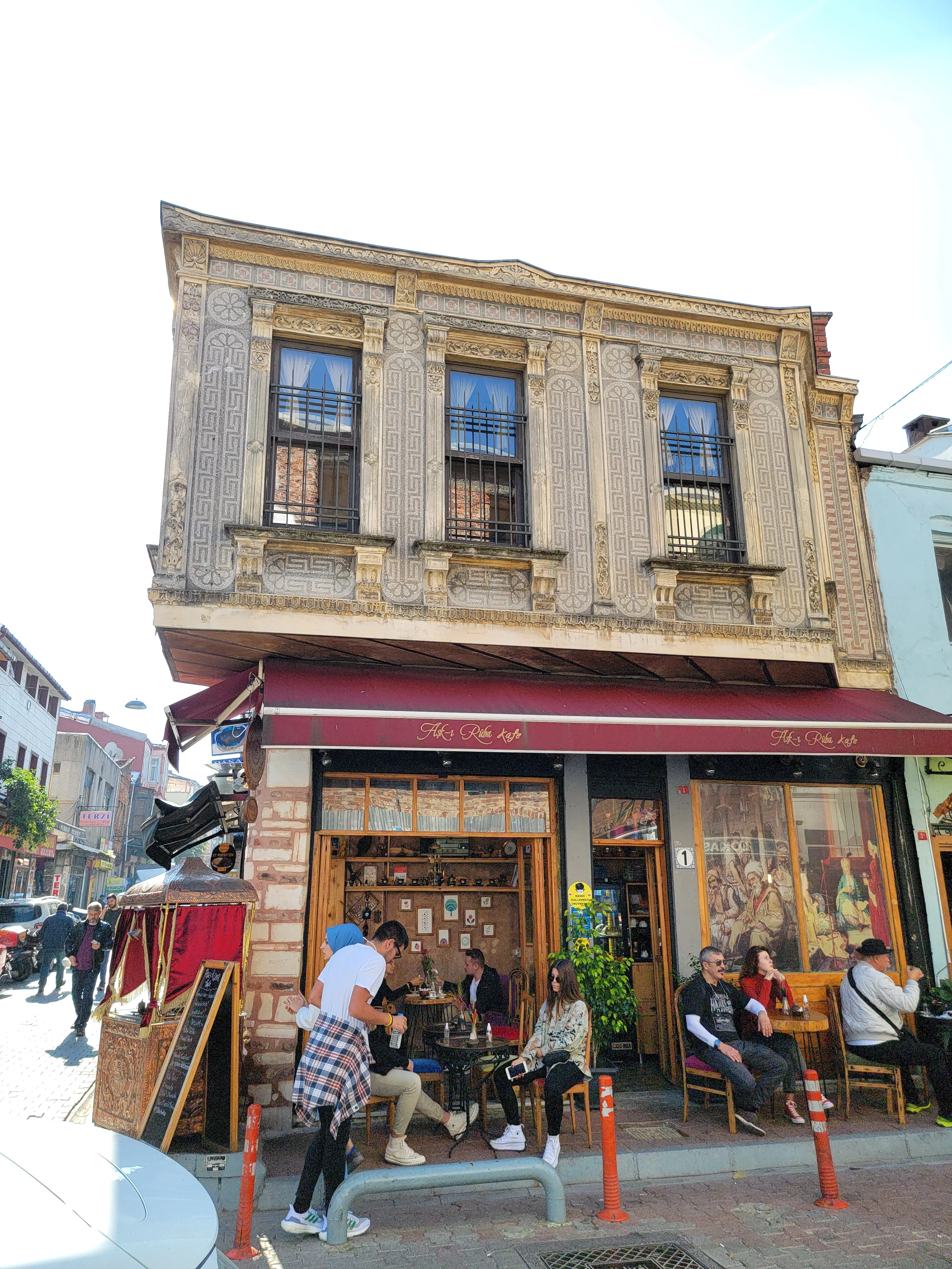 A two-story building with a rustic facade houses a café. People sit and chat at tables outside under a red awning, while others walk by on the street. Sunlight brightens the scene.