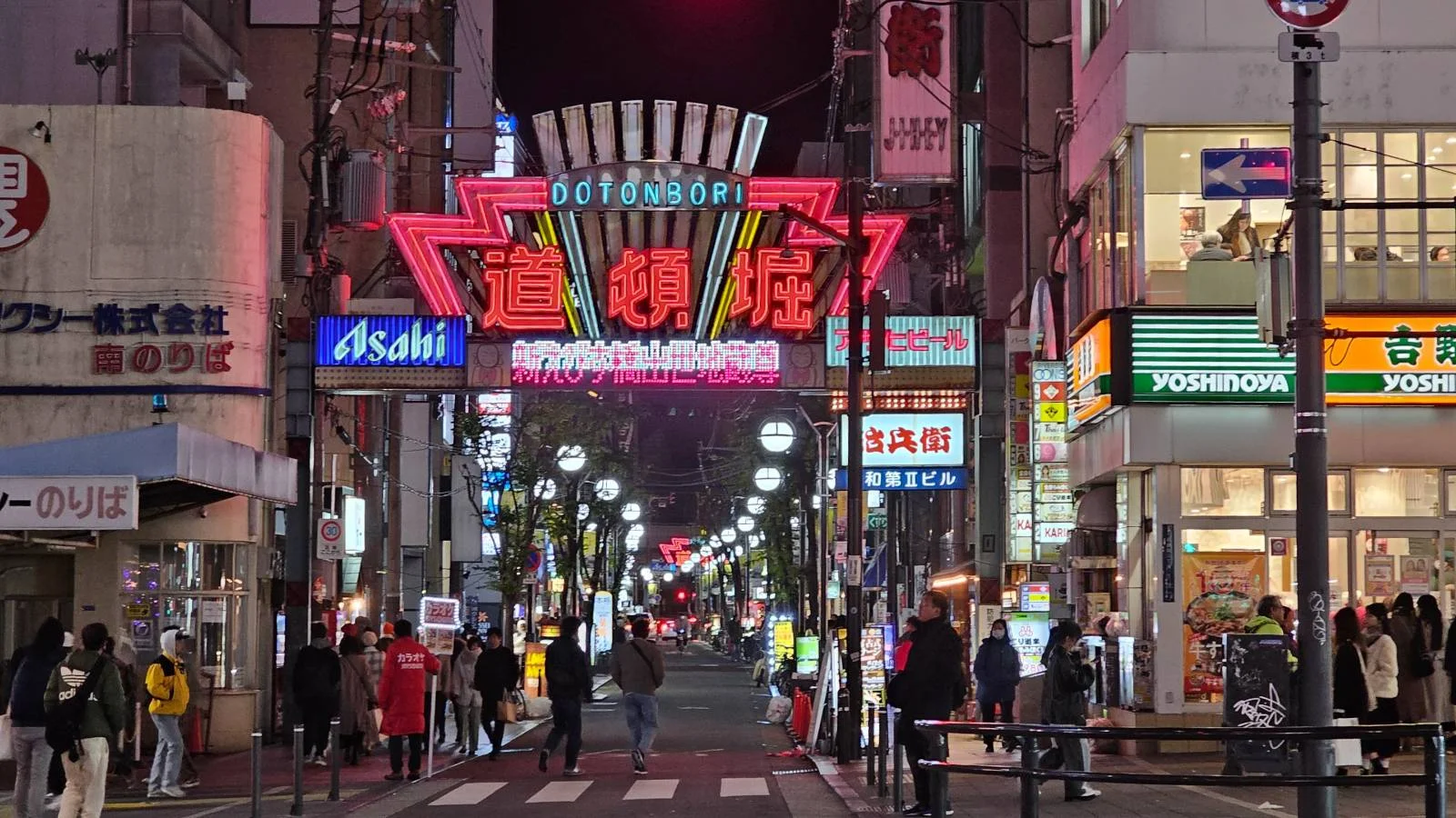 Night scene in Dotonbori, Osaka with neon signs and a bustling crowd. Notable signs: Dotonbori, Asahi, Yoshinoya. Lively urban vibe.