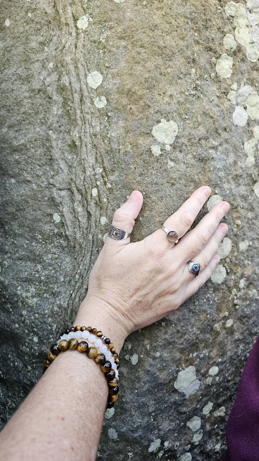 A hand wearing several rings and beaded bracelets touches the surface of a large, textured rock with small patches of lichen.