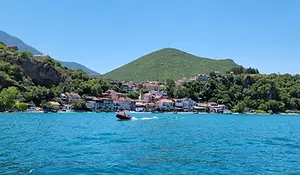Village along Lake Ohrid with red-roofed houses set against green hills, viewed from the water with a small boat crossing the lake.