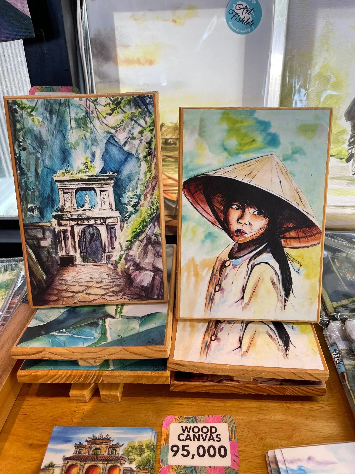 Two paintings on a display stand. The left painting depicts an ancient stone gate surrounded by greenery. The right painting features a person wearing a traditional conical hat and robe, looking back with a smile. Price tags are visible below.