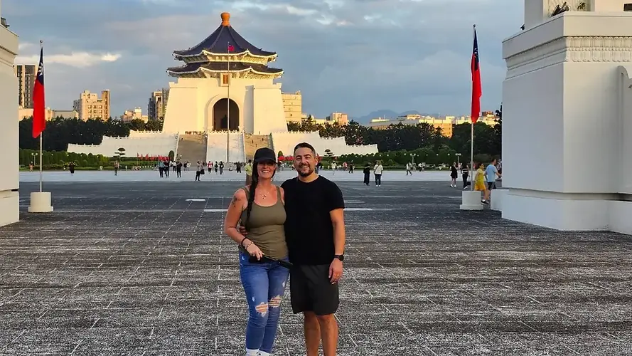 Jenn and Leon smiling in the expansive plaza in front of the iconic white and blue Chiang Kai-shek Memorial Hall in Taipei, Taiwan, representing their global travel lifestyle.