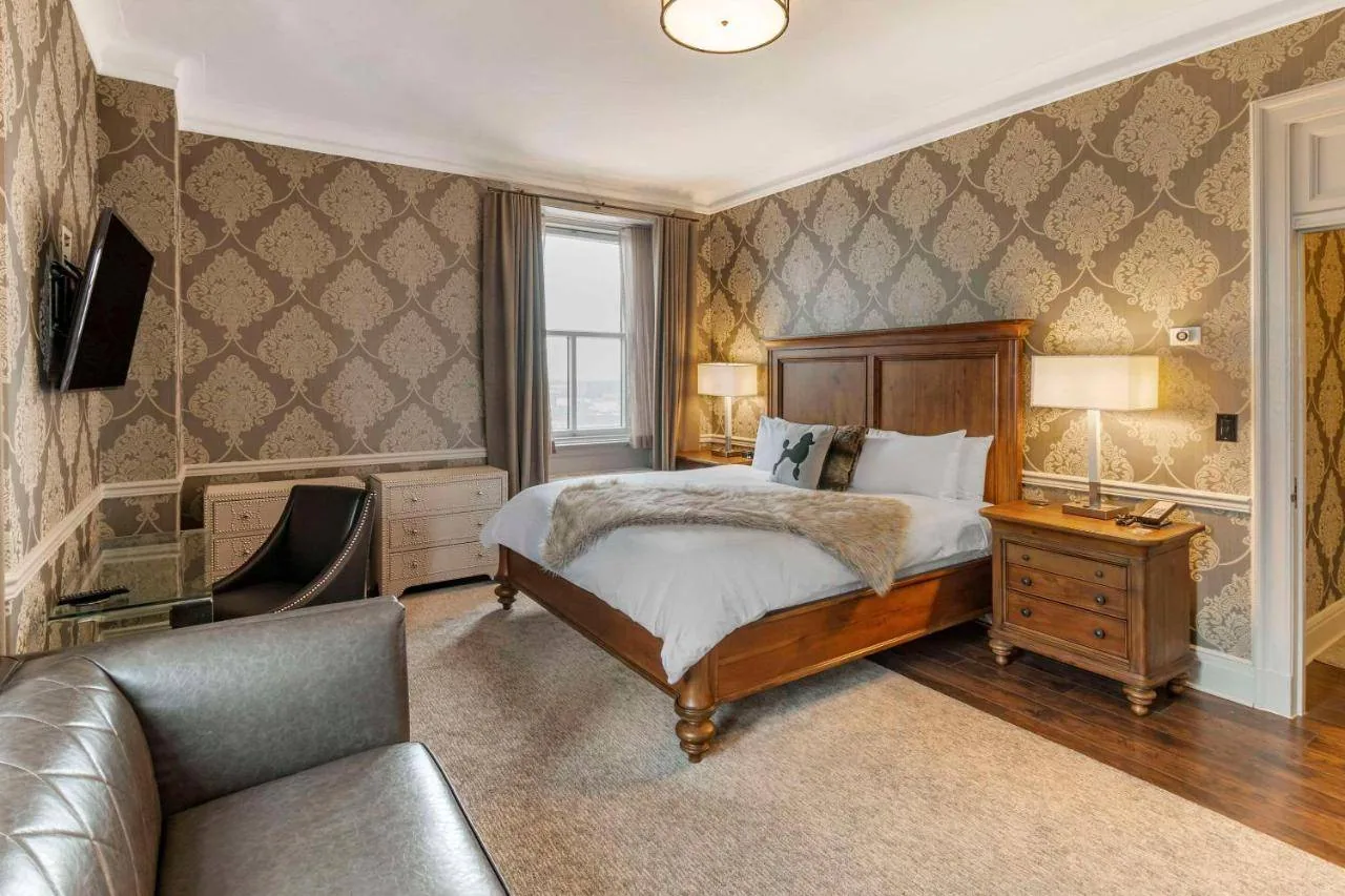 A cozy hotel room with a large bed featuring white linens and a throw blanket. The walls are decorated with patterned wallpaper. There's a TV mounted on one wall, a leather chair, a desk, and two bedside tables with lamps. A window lets in natural light.