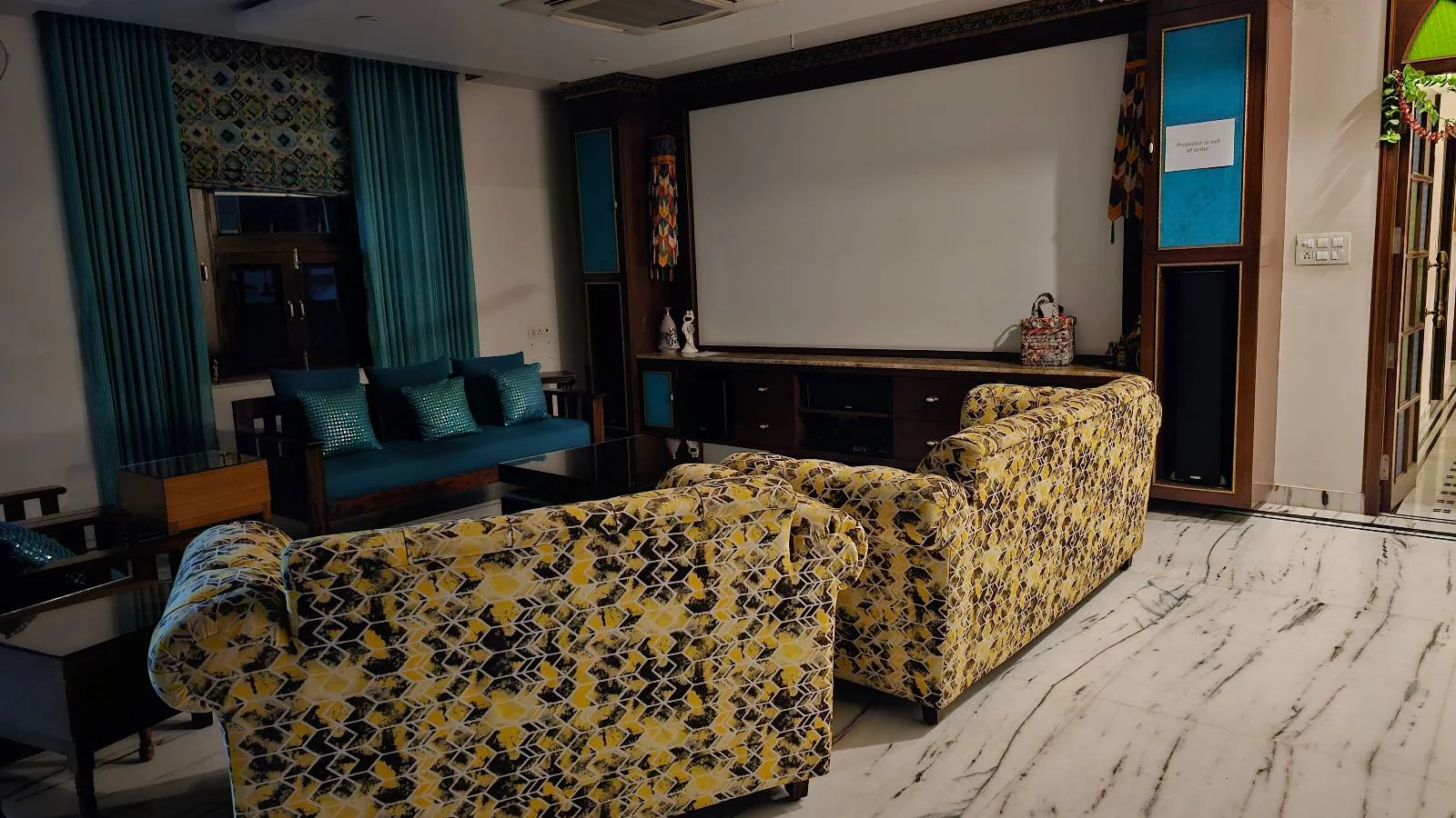 A cozy home theater room with floral-patterned couches facing a large projector screen, blue-accented walls, and a small sitting area with teal cushions near a window.