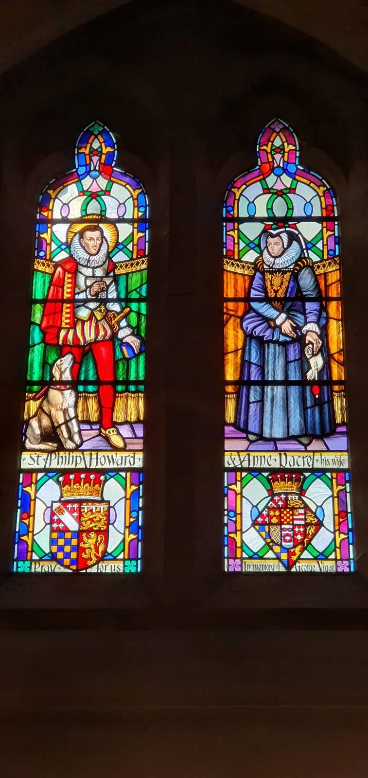 Stained glass window depicting two figures in colorful attire. The left figure is in armor with a shield and sword, accompanied by a dog. The right figure is in a blue robe, holding a book. Both panels are ornately decorated with intricate patterns.