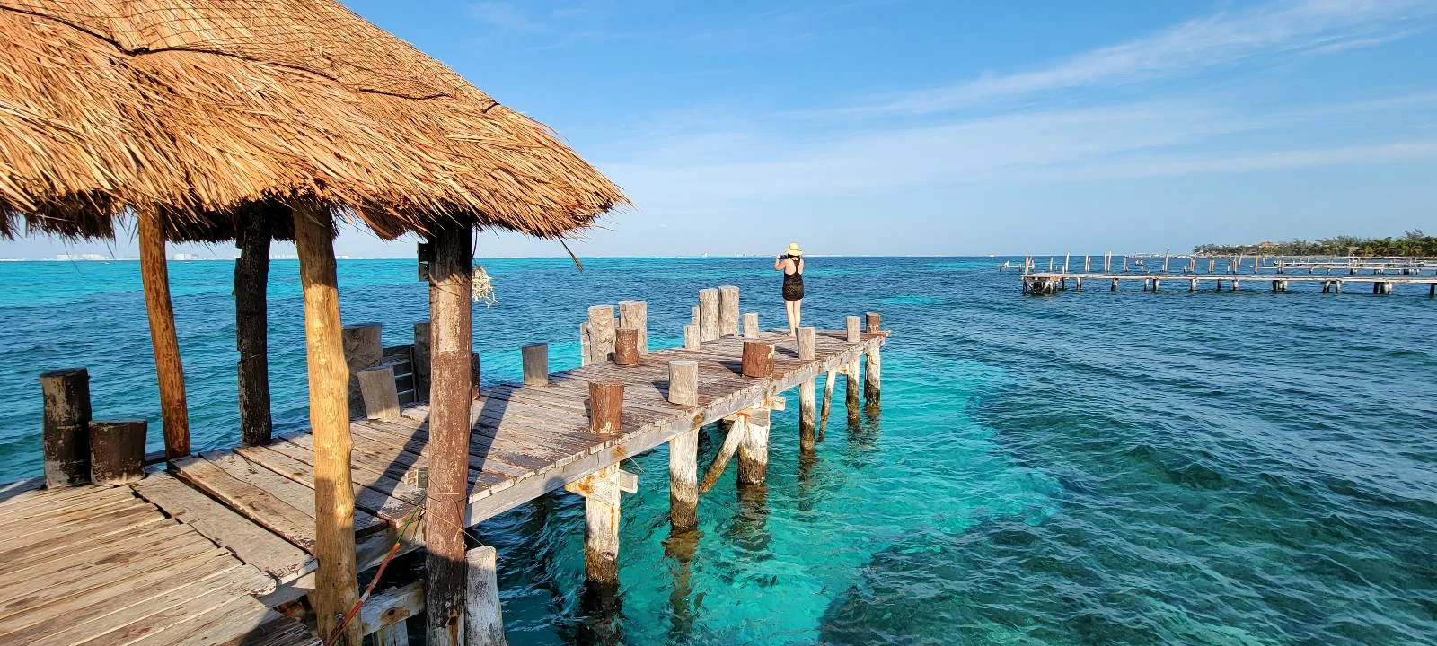 A wooden pier extends into clear turquoise water, surrounded by a thatched roof structure on the left. The sky is bright blue with a few clouds, creating a serene and tropical atmosphere.