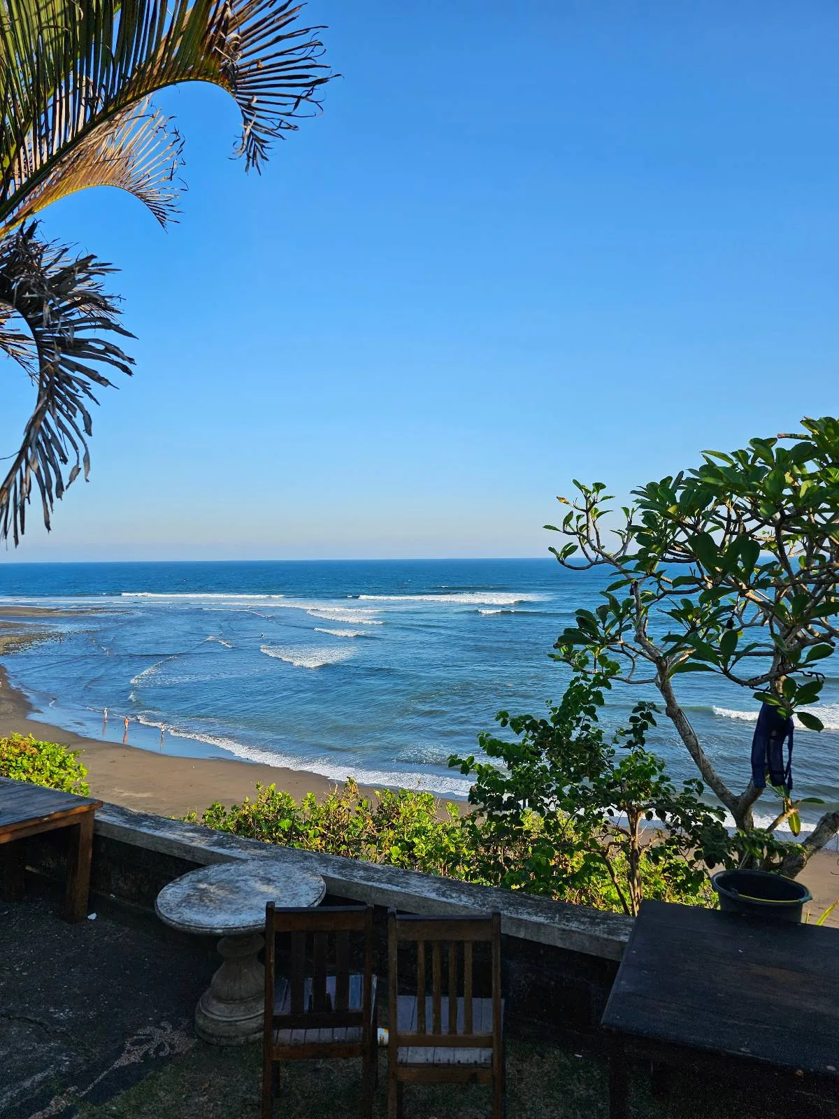 A tranquil beach scene with clear blue skies and gentle ocean waves. Two wooden chairs and a small stone table overlook the water, surrounded by palm trees and lush greenery.
