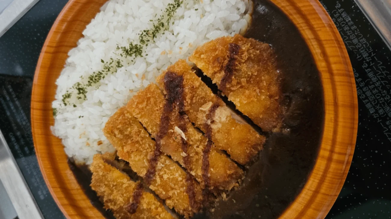 A bowl of Japanese curry with steamed white rice and sliced breaded fried pork cutlet (katsu), garnished with green herbs on top of the rice.