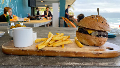 A burger with cheese and fries on a wooden board, accompanied by a white cup, is placed on a gray table. In the background, people are seated at tables on a covered patio overlooking a beach.