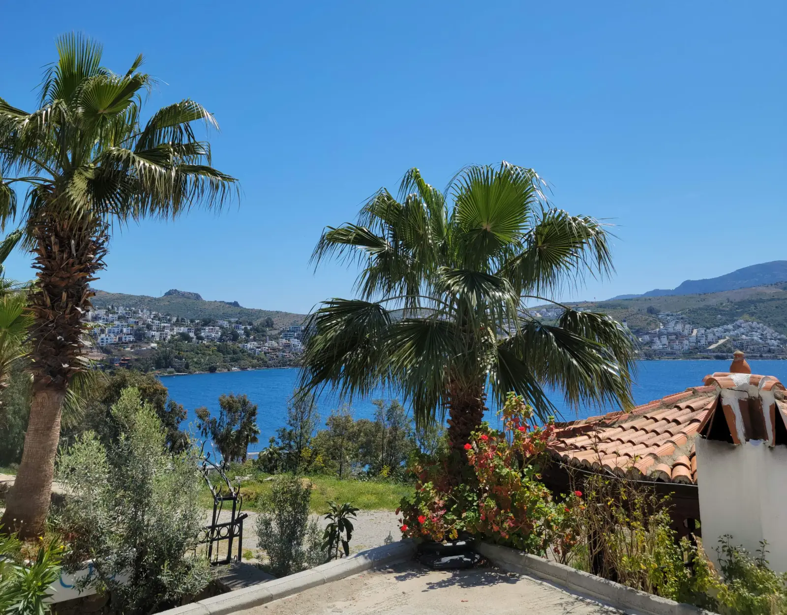 View of a coastal landscape featuring palm trees, a calm blue sea, distant hills, and a tiled-roof house on a sunny day.
