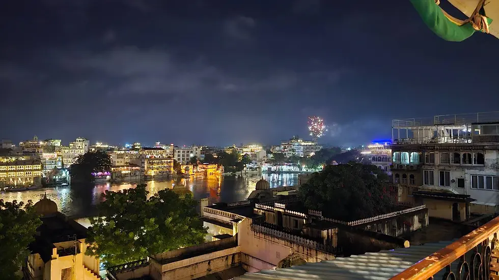 Night cityscape with brightly lit riverside buildings. Fireworks in the sky create a festive atmosphere. Reflective water enhances the scene.