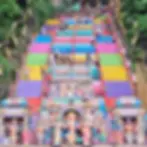 Vibrant, multicolored steps lead up to Batu Caves, surrounded by lush greenery. Statues of Hindu deities adorn the staircase and entrance, with visitors ascending the steps.