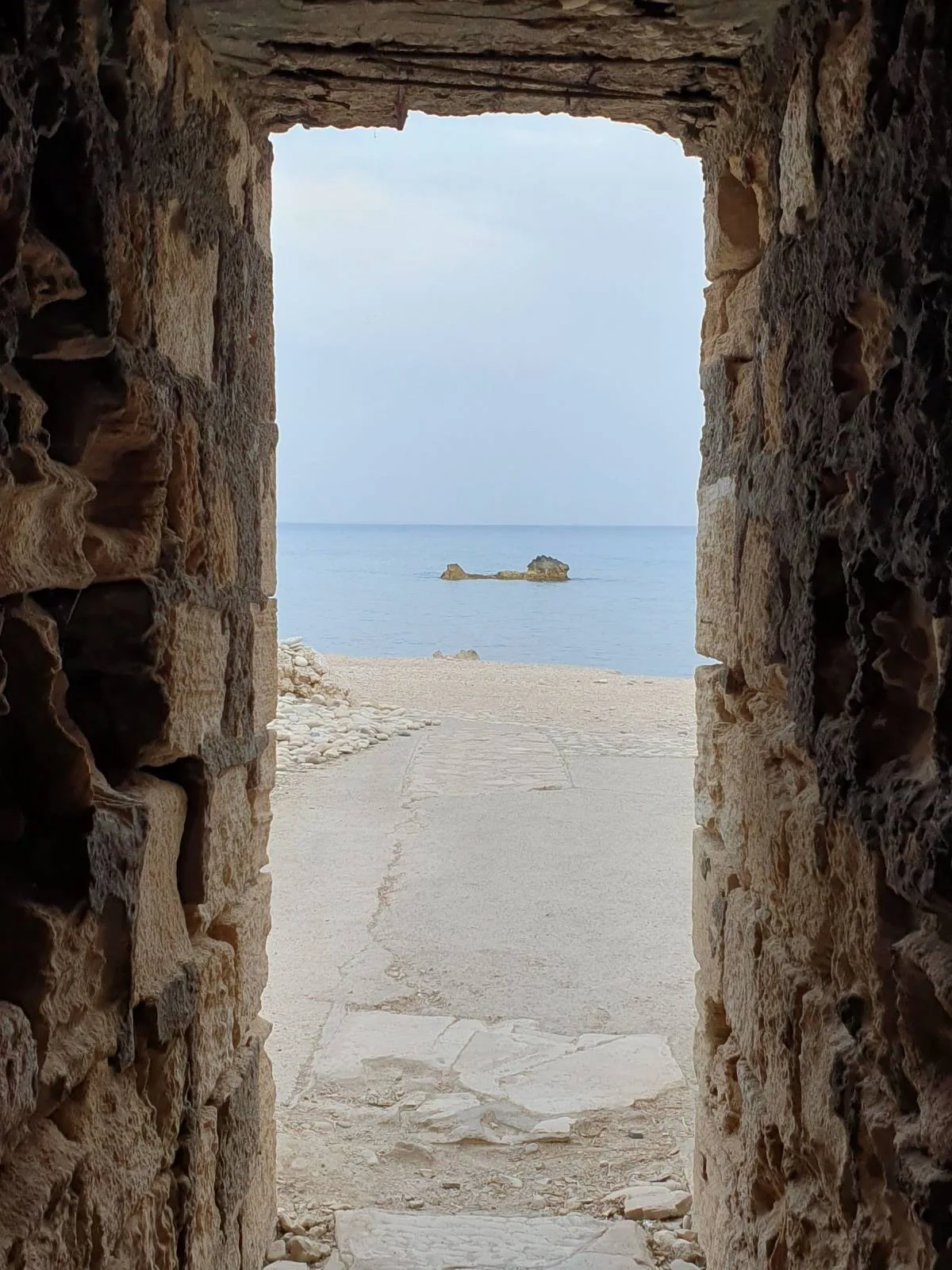 View through a stone archway leading to a sandy beach with a calm blue sea in the background. A small rocky formation is visible in the water under a cloudy sky.
