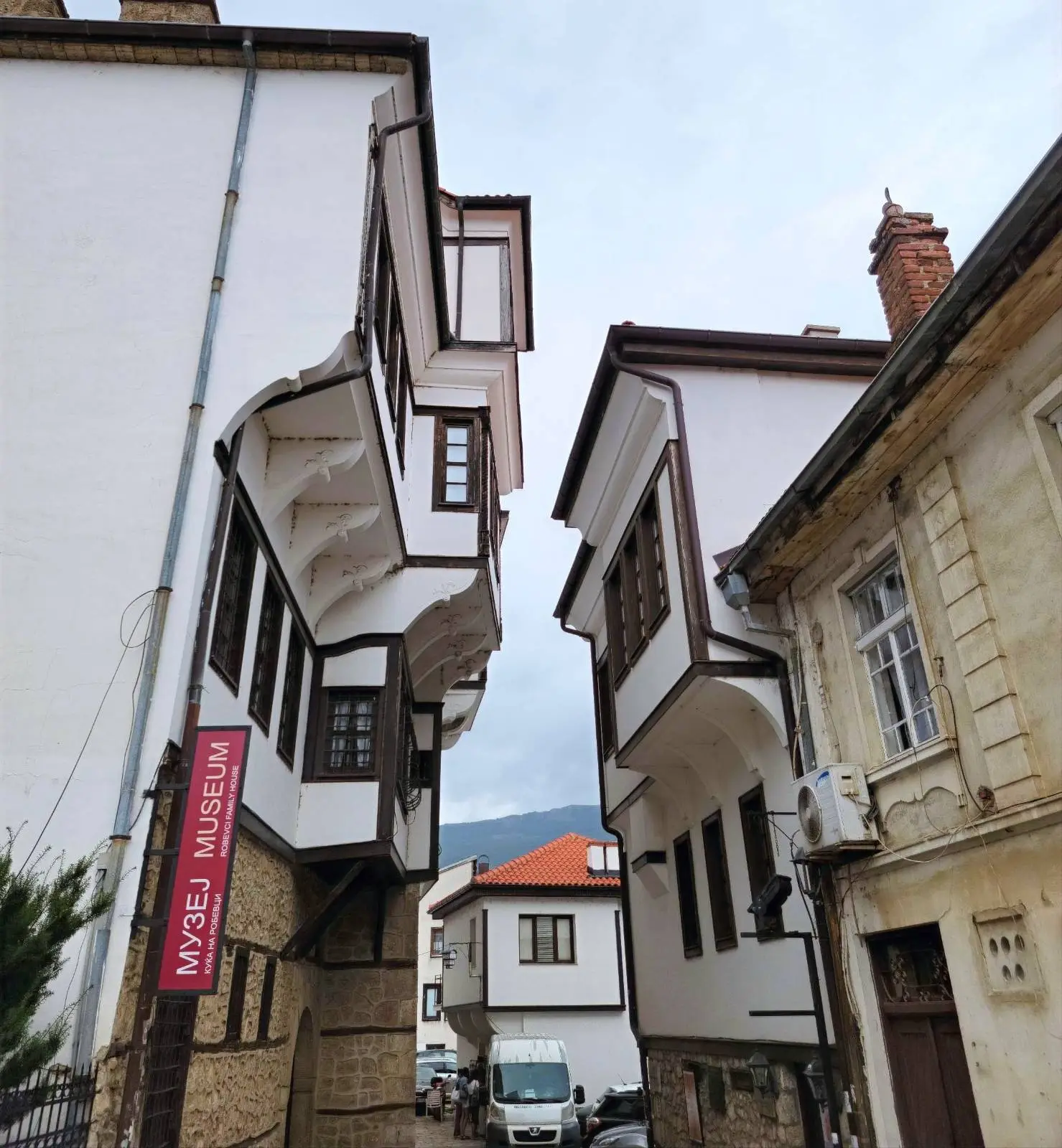 Two buildings with traditional architecture nearly touch at the top, forming a narrow passageway. The building on the left has a museum sign. A small white truck is parked below, with a cloudy sky overhead.