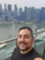 Smiling man takes a selfie on a balcony with a city skyline backdrop. Skyscrapers and water visible, creating a bright, urban scene.