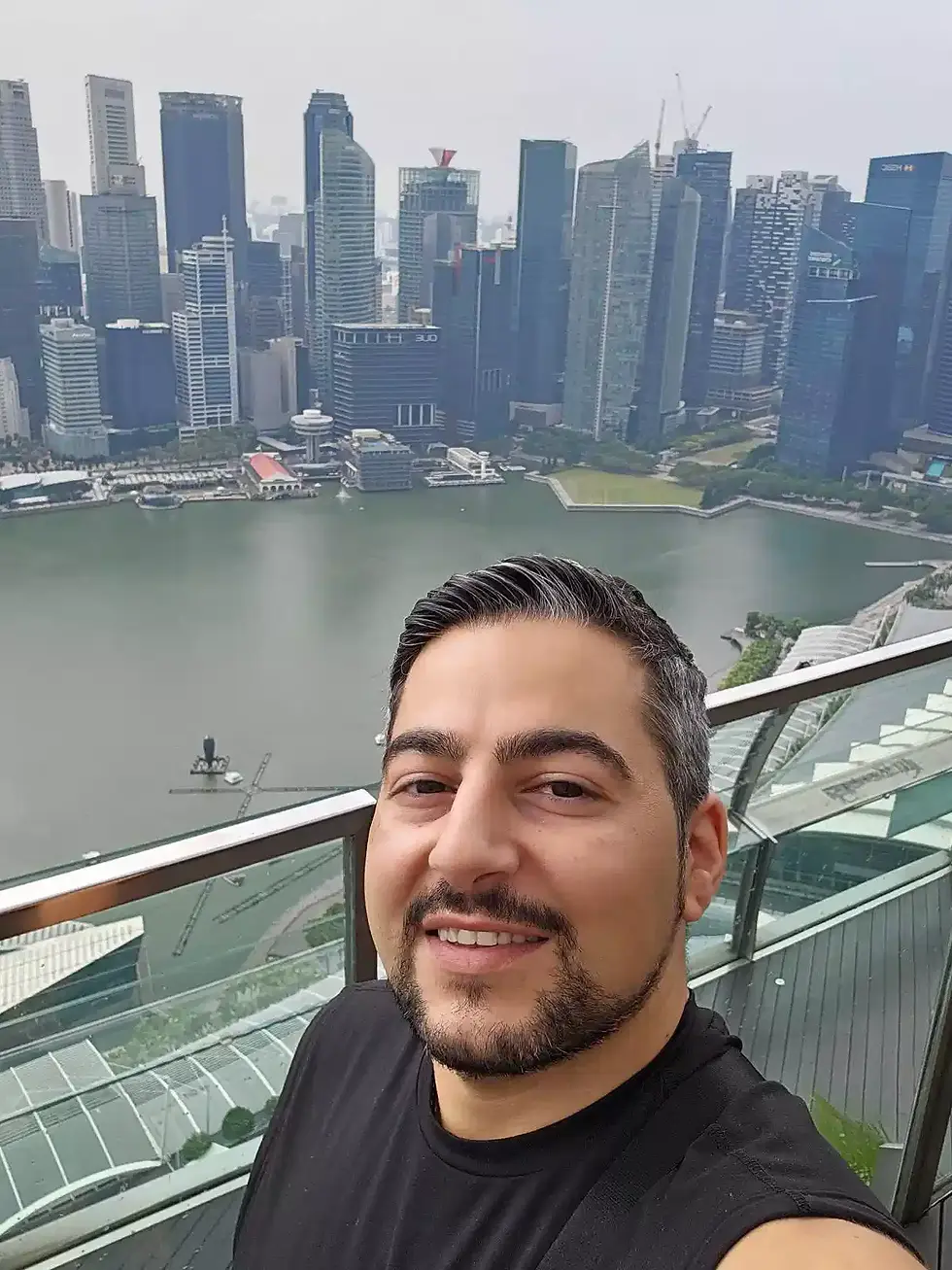 Smiling man takes a selfie on a balcony with a city skyline backdrop. Skyscrapers and water visible, creating a bright, urban scene.