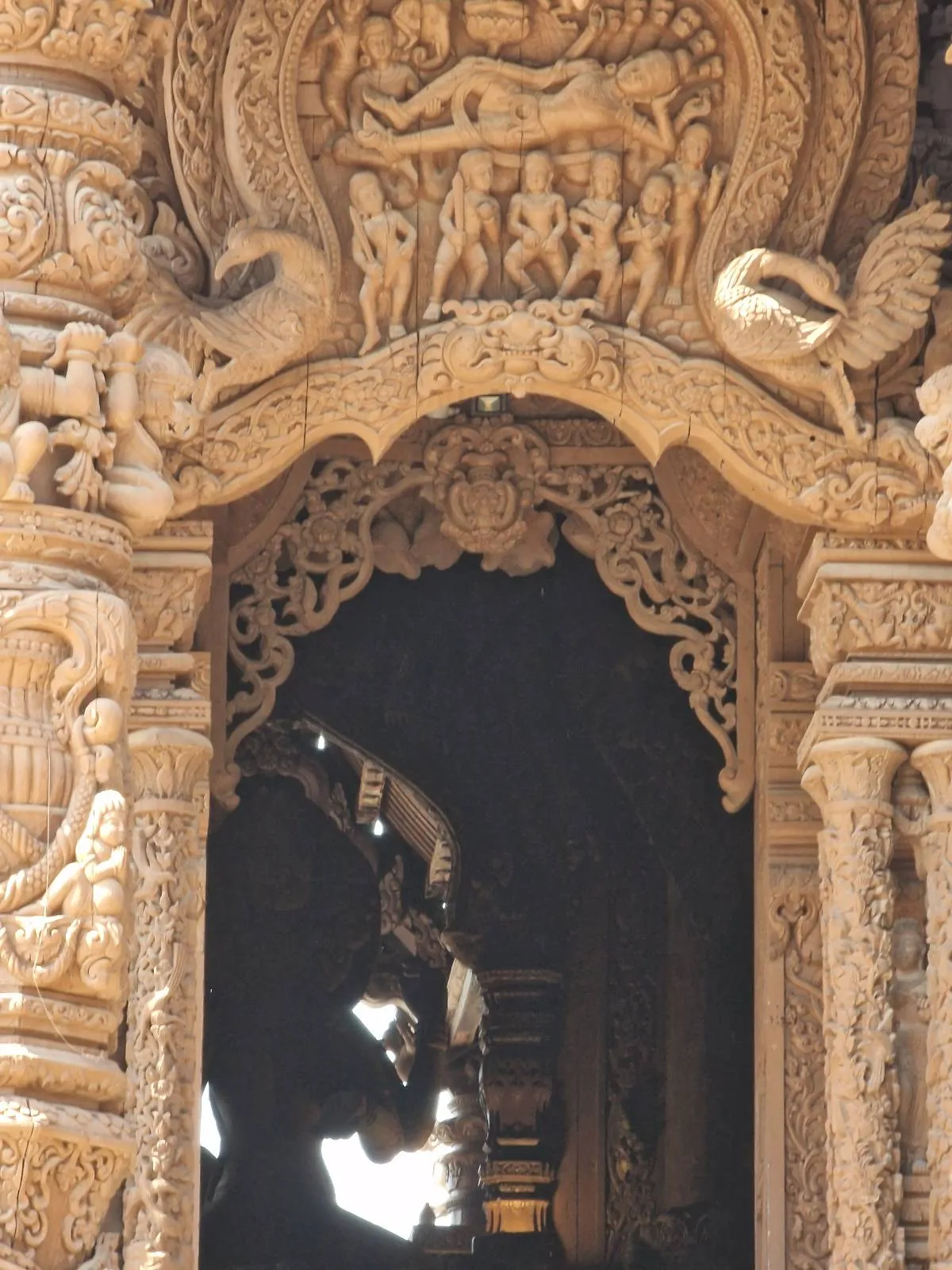 Intricately carved wooden archway adorned with mythical creatures, human figures, and ornate patterns. The detailed craftsmanship highlights artistic elements and cultural symbols. Soft light casts shadows, enhancing the texture of the carvings.