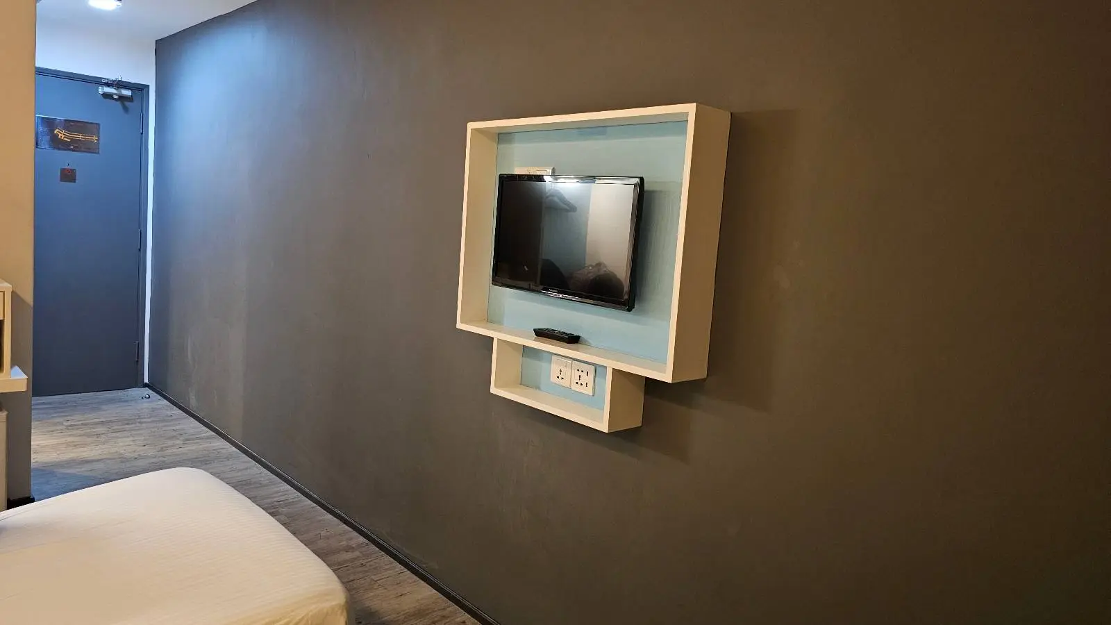 A flat-screen TV is mounted on a dark gray wall in a minimalist room, framed by a light-colored rectangular border. A small shelf beneath the TV holds a digital clock.